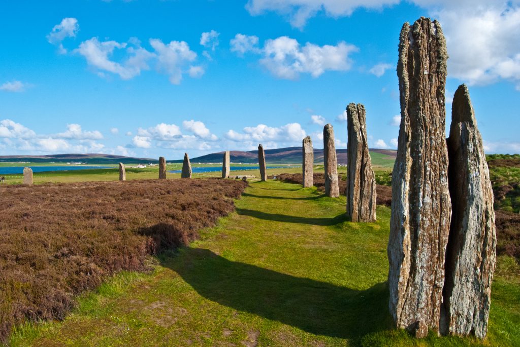 Ein beeindruckender Kreis aus aufrechten, alten Steinen steht in einer weiten, hügeligen Landschaft unter blauem Himmel.