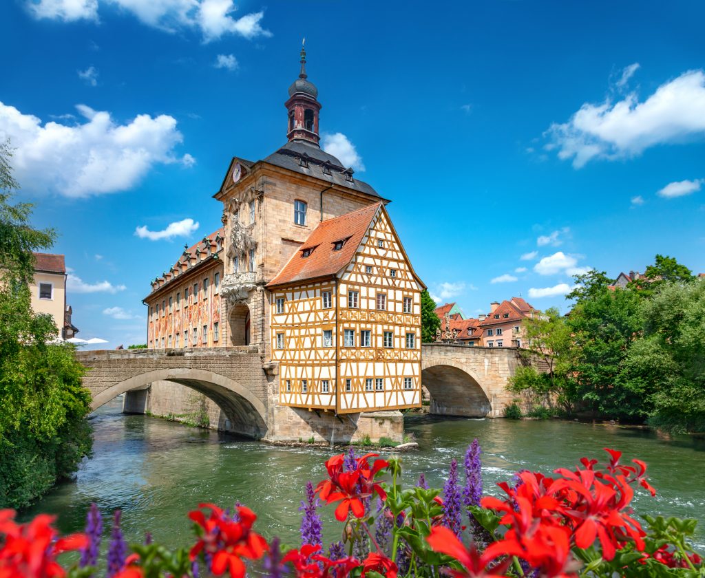 Rote Geranien blühen im Vordergrund vor dem berühmten Rathausgebäude auf der Brücke über die Regnitz.