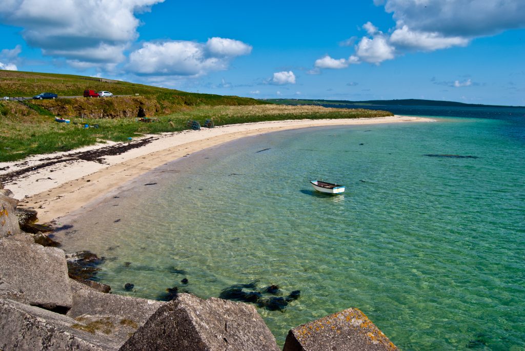 Ein kleines Ruderboot liegt in kristallklarem Wasser vor einem hellen Sandstrand an der Küste von Orkney.