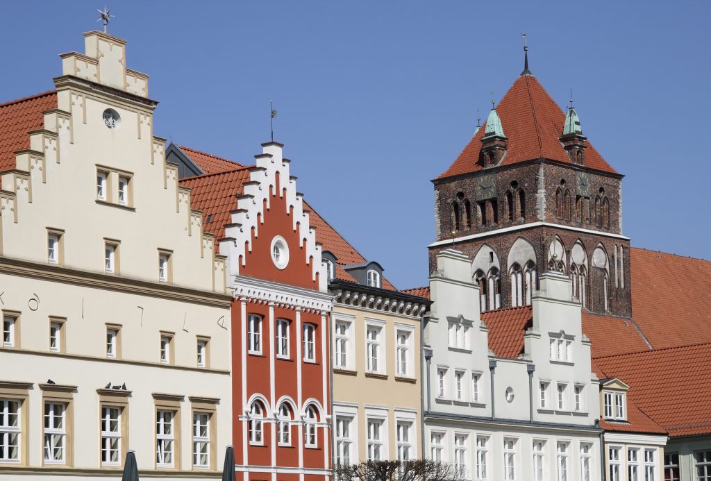 Eine Reihe historischer Giebelhäuser in verschiedenen Farben steht neben einem alten Backsteinkirchturm unter strahlend blauem Himmel.