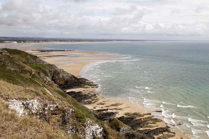Ein weiter Blick über eine felsige Küste und einen langen Sandstrand bei Cherbourg unter wolkigem Himmel.