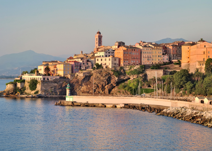 Das Licht der untergehenden Sonne beleuchtet die Fassaden der Altstadt von Bastia und das ruhige Wasser im Hafenbecken.