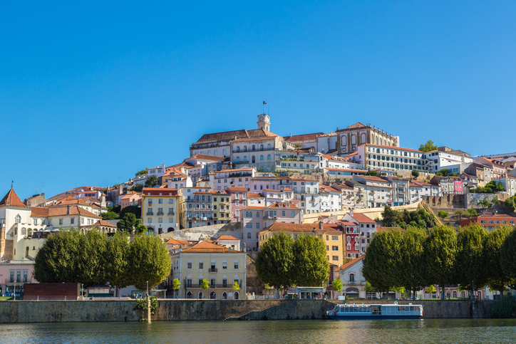 Die malerische Altstadt von Coimbra erstreckt sich am Ufer des Flusses Mondego mit historischen Gebäuden und einem Kirchturm.