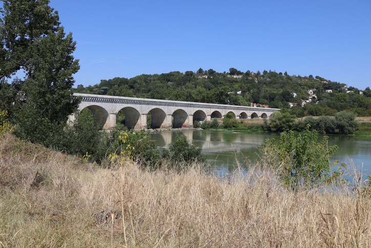 Eine historische Steinbrücke mit vielen Bögen überspannt einen ruhigen Flussabschnitt in der Nähe von Agen.