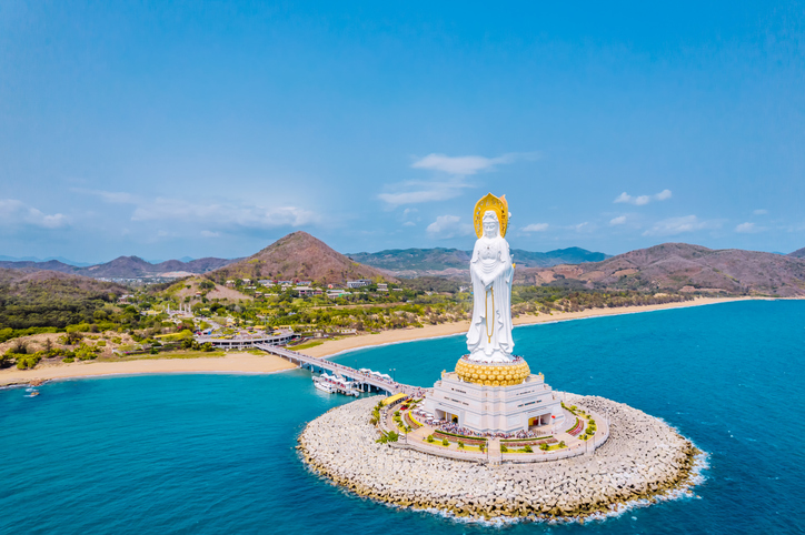 Eine riesige, weiße Statue der Gottheit Guanyin steht auf einer künstlichen Insel im Meer vor der Küste von Hainan, verbunden durch eine lange Brücke.