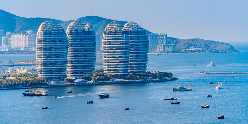 Futuristisch geformte Hochhäuser ragen direkt am Ufer einer von Booten befahrenen Bucht in Hainan in den Himmel.