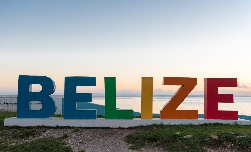 Ein großer, bunt bemalter Schriftzug mit dem Wort „BELIZE“ steht an einer Küste vor einem ruhigen Meer während des Sonnenuntergangs.