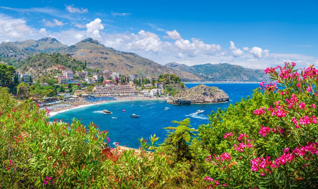 Blick von oben auf das tiefblaue Meer und die Bucht von Taormina, eingerahmt von blühenden Oleandersträuchern