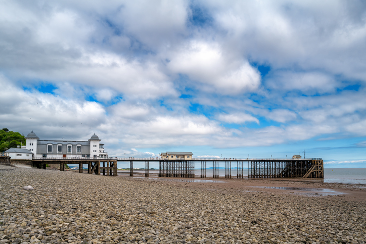 Ein langer Pier führt über den Kiesstrand hinaus aufs Meer unter einem wolkigen Himmel.