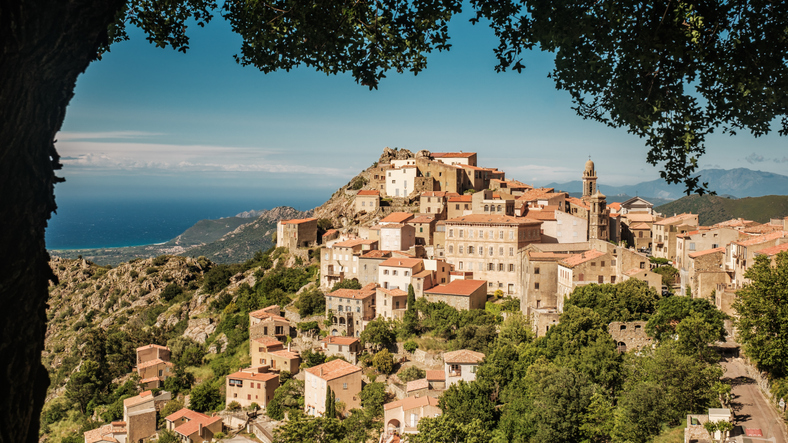 Ein auf einem Hügel erbautes Dorf mit eng stehenden Steinhäusern, umrahmt von mediterraner Vegetation auf Korsika.