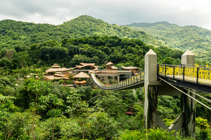 Eine schmale Hängebrücke führt durch ein üppig grünes Waldgebiet in den Bergen von Hainan zu einem Gebäude mit traditionellen Dächern.