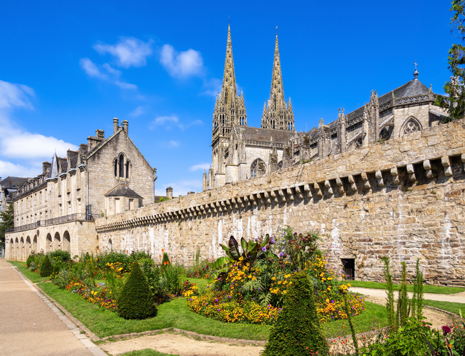 Ein prachtvoller Blumengarten im Vordergrund, dahinter eine hohe Steinmauer und die imposanten Türme der Kathedrale von Quimper
