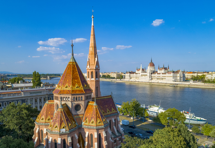 Eine Kirche mit buntem Dach steht am Donauufer, im Hintergrund ist das ungarische Parlament zu sehen.