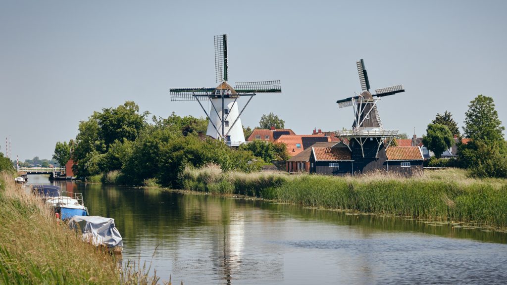 Zwei Windmühlen stehen in einer grünen, ruhigen Landschaft direkt an einem kleinen Kanal.
