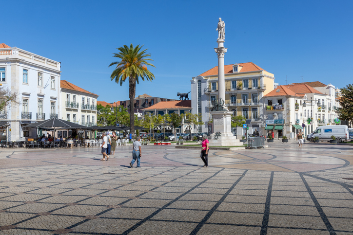 Ein hoher weißer Obelisk mit einer Statue auf einem weitläufigen Platz, umgeben von traditionellen Gebäuden mit Dachterrakotta-Ziegeln