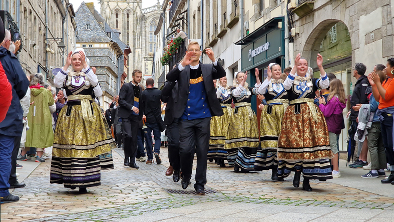 Männer und Frauen in traditioneller bretonischer Tracht gehen in einer Prozession durch eine enge, kopfsteingepflasterte Gasse, gesäumt von historischen Gebäuden