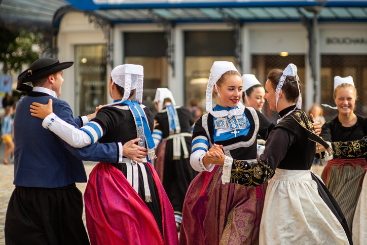 Eine Gruppe von Menschen in prachtvollen, historischen bretonischen Trachten mit weißen Hauben tanzt gemeinsam auf einem öffentlichen Platz