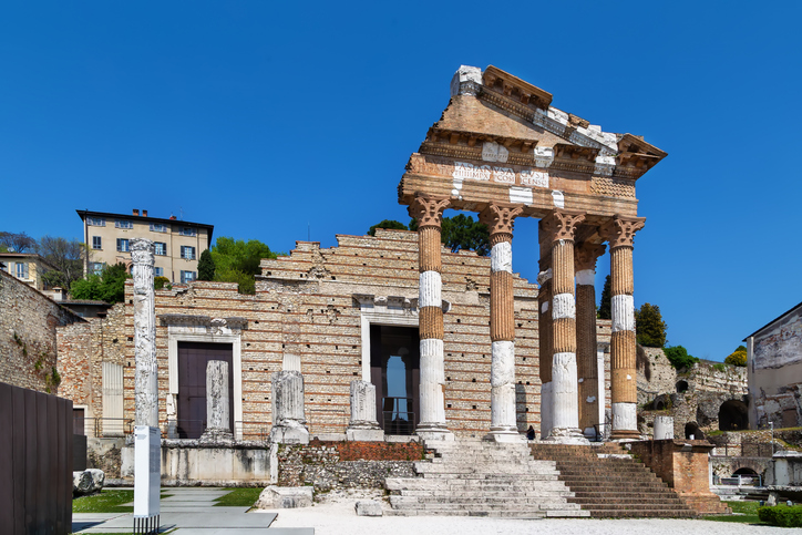 Die antike Tempelfassade des Capitolium mit ihren Säulen und dem steinernen Treppenaufgang vor klarem blauem Himmel.
