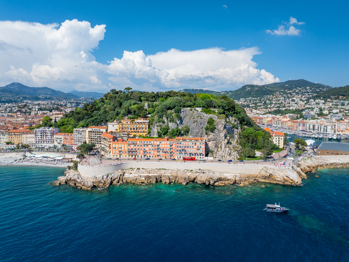 Blick auf die Küste von Nizza mit Felsen, Meer, Stadt und grüner Anhöhe.