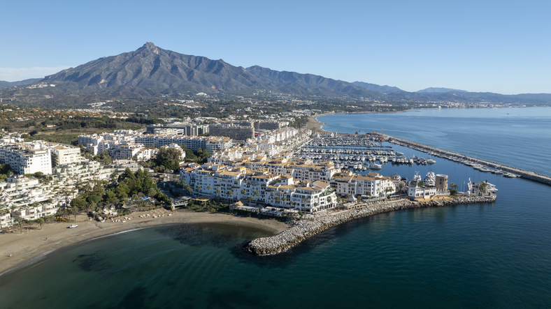 Eine Luftaufnahme zeigt den modernen Yachthafen von Marbella mit zahlreichen Booten, angrenzenden Gebäuden und einem Sandstrand direkt am Mittelmeer.