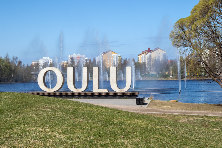 Die großen, weißen Buchstaben „OULU“ stehen auf einem Podest an einem See mit Wasserfontänen im Hintergrund.