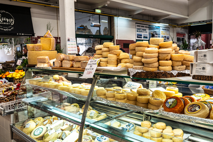 Eine gut gefüllte Kühltheke mit verschiedenen Käselaiben in einer Markthalle, im Hintergrund sind Regale mit regionalen Produkten sichtbar