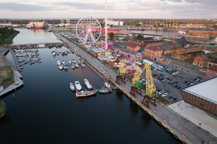 Ein belebter Hafenbereich aus der Vogelperspektive mit Riesenrad, Fahrgeschäften und kleinen Booten im Wasser.