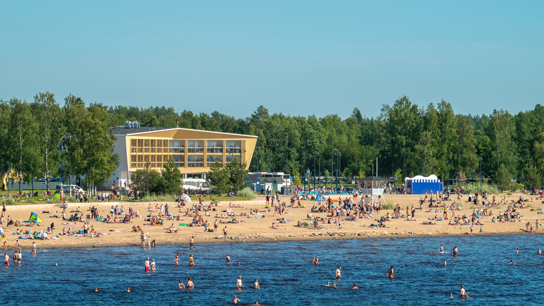 Ein belebter Sandstrand mit vielen Menschen, die bei strahlendem Wetter schwimmen oder entspannen.