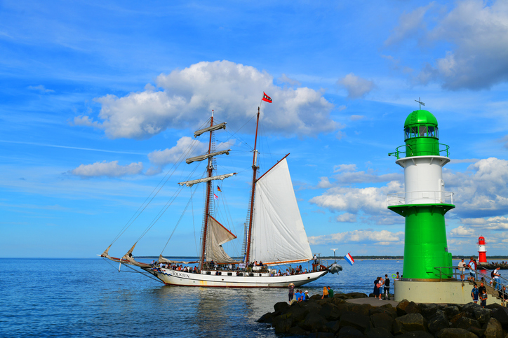 Ein großes Segelschiff fährt an der markanten Hafeneinfahrt von Warnemünde mit dem grünen Leuchtturm vorbei.