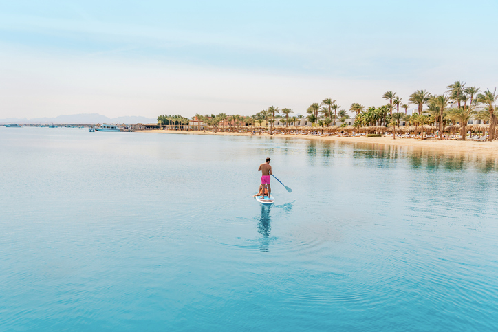 Ein Mann fährt auf einem Stand-Up-Paddle-Board durch das spiegelglatte Wasser vor einer Hotelanlage in Hurghada.