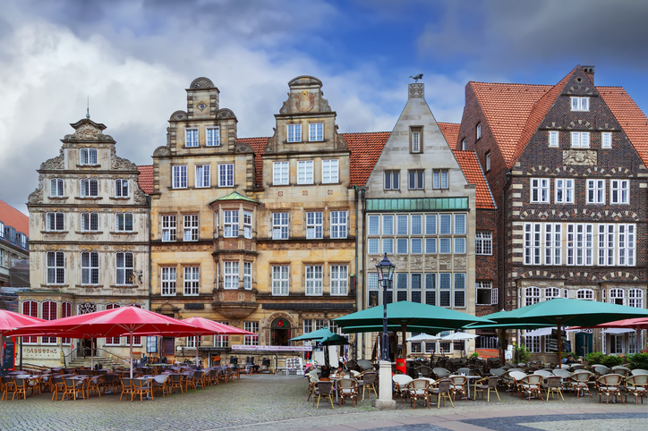 Eine Reihe von prunkvollen, historischen Giebelhäusern säumt den Bremer Marktplatz, während davor Menschen an kleinen Tischen unter großen Sonnenschirmen sitzen.