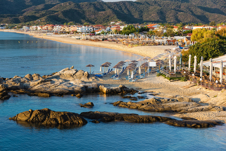 Blick auf einen Strandabschnitt in Sarti mit großen Felsen im Vordergrund und Sonnenschirmen im Hintergrund.