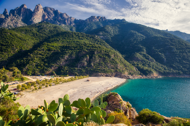 Ein weiter Sandstrand mit einem kleinen Gebäude, eingebettet zwischen einer grünen Bergkette und dem tiefblauen Meer auf Korsika.