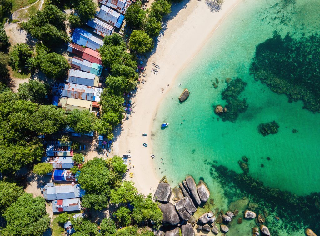 Vogelperspektive auf einen Sandstrand mit Booten und bunten Hütten, eingebettet zwischen dichtem Grün und türkisfarbenem Meer.