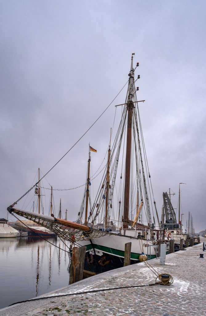 Ein historisches Segelschiff mit beeindruckendem Mast liegt an einer steinernen Kaimauer in einem ruhigen Hafen.