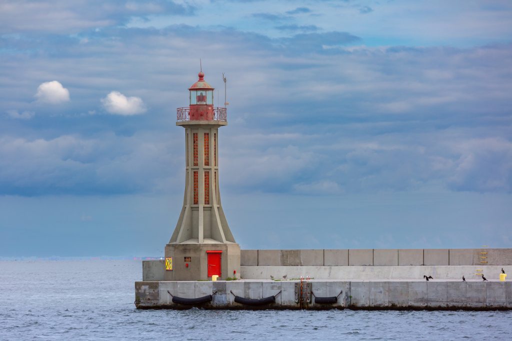 Ein schmaler, grauer Leuchtturm mit roter Laterne steht einsam am Ende einer betonierten Kaimauer im Meer.