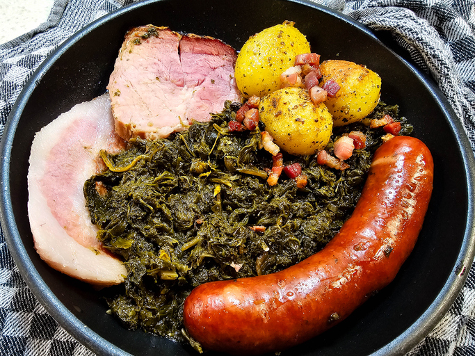 Ein schwarzer Teller mit Grünkohl, einer deftigen Wurst und Kasseler sowie kleinen Kartoffeln steht auf einem karierten Tuch.