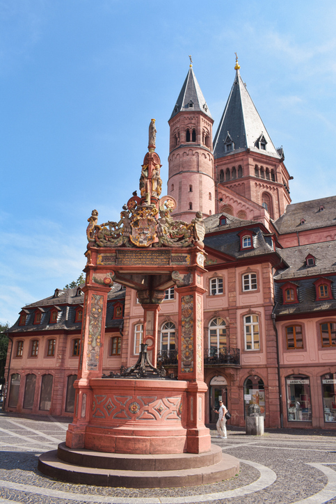 Ein kunstvoll verzierter, roter Sandsteinbrunnen steht auf einem leeren Platz vor den hohen Türmen des Mainzer Doms.