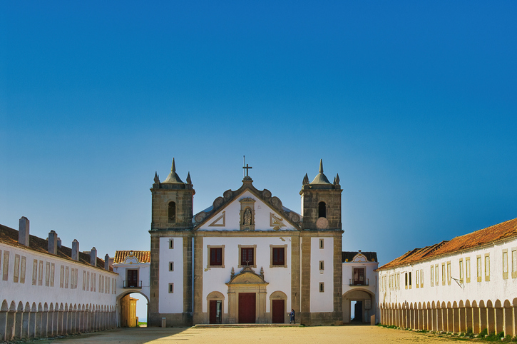 Eine imposante, weiße Kirche mit zwei Türmen vor einem strahlend blauen Himmel, umgeben von symmetrischen Seitengebäuden mit Bogengängen