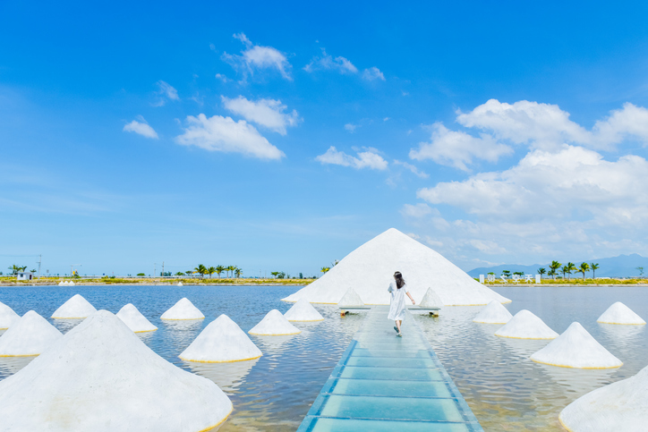 Eine Person spaziert über einen Glassteg auf einer Salzfarm in Hainan, umgeben von weißen Salzpyramiden in flachem Wasser unter blauem Himmel.