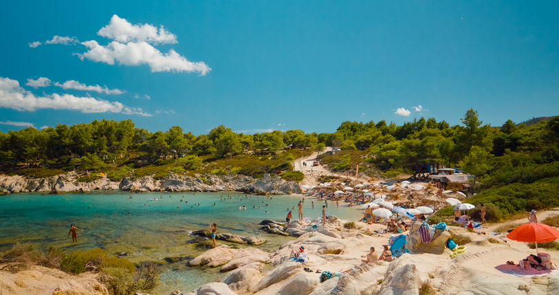 Ein lebendiger Strand in Sarti, an dem Menschen zwischen Felsen im kristallklaren Wasser schwimmen und sonnenbaden.