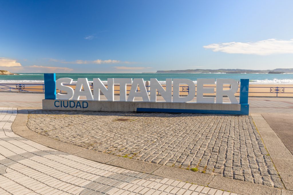 Ein weißer, großer „SANTANDER“-Schriftzug steht auf einer gepflasterten Promenade direkt am Meer.