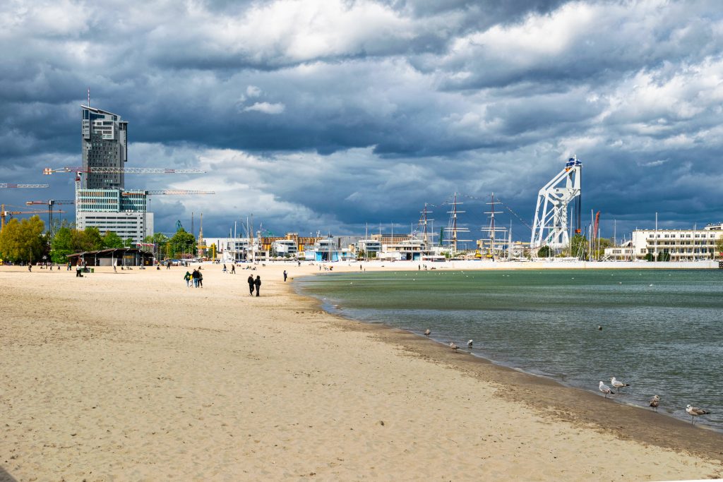 Ein weitläufiger Sandstrand unter bewölktem Himmel mit Blick auf den modernen Gdynia-Hafen und markante Hochhäuser.