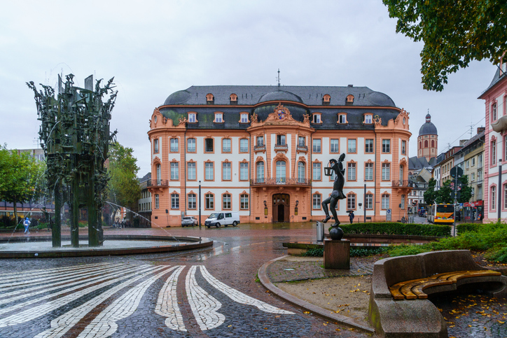 Ein prachtvoller Bau mit pfirsichfarbener Fassade steht an einem regnerischen Platz in Mainz, auf dem ein skurriler Brunnen und Skulpturen zu sehen sind.