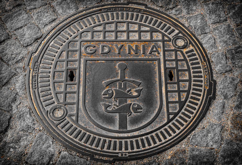Ein gusseiserner Kanaldeckel auf Kopfsteinpflaster, verziert mit dem Wappen von Gdynia, das ein Schwert und zwei Fische zeigt.