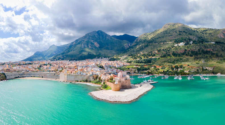 Ein Luftbild von Trapani, das die Stadt auf einer Halbinsel zwischen tiefblauem Meer und dem Bergland im Hintergrund zeigt.