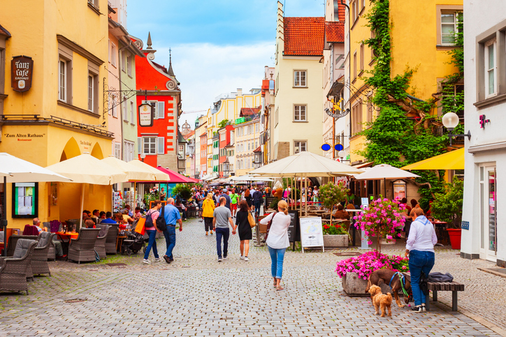 Eine belebte, gepflasterte Gasse mit historischen, bunten Giebelhäusern, zahlreichen Straßencafés unter Sonnenschirmen und Menschen, die den Tag genießen
