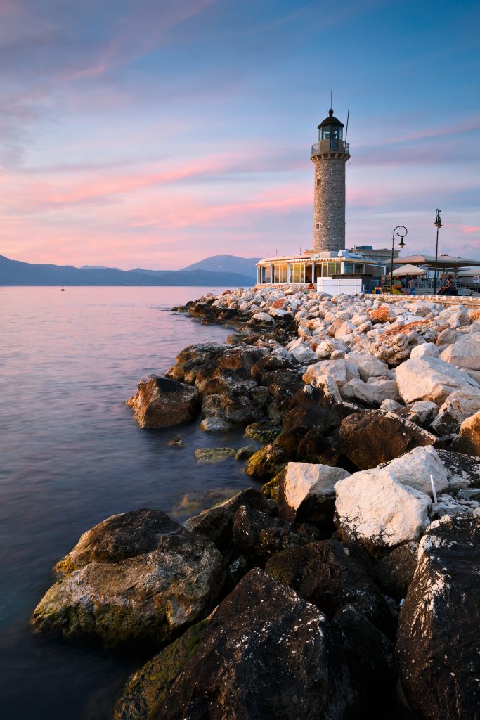 Ein steinerner Leuchtturm steht an einer felsigen Küste während eines rosafarbenen Sonnenuntergangs.