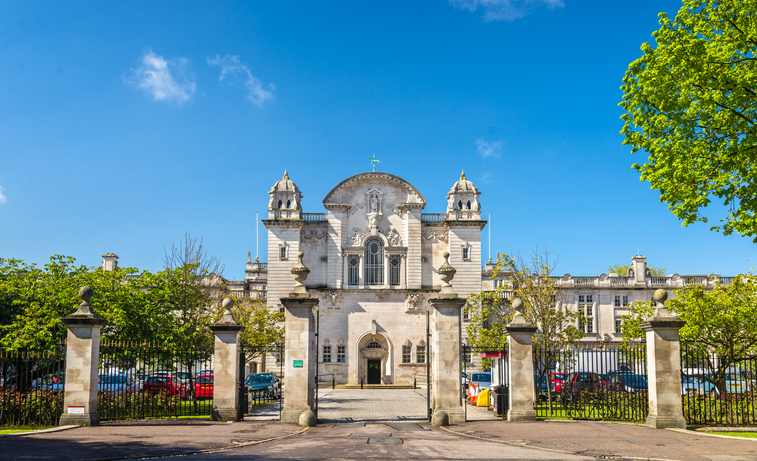 Ein helles historisches Gebäude steht hinter einem Zaun im grünen Stadtzentrum von Cardiff.