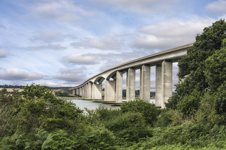 Eine mächtige Betonbrücke spannt sich über das Mündungsgebiet des Flusses, umgeben von grüner Natur.